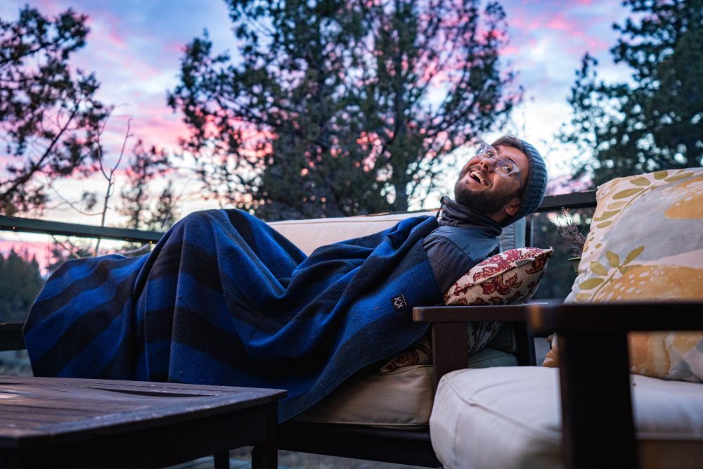 Person lying on a couch under a blue Arcturus blanket with a scenic background of trees and sky.